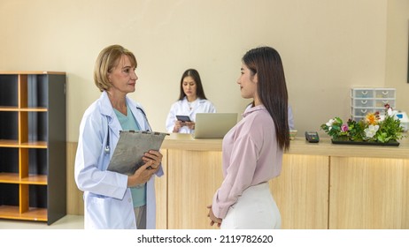 Elderly Blonde Hair Surgical Doctor Discusses With Her Patient For A Cosmetic Or Beauty Treatment Program At Reception Desk. Female Girl Gets Suggestion From A Female Doctor In A Beauty Clinic