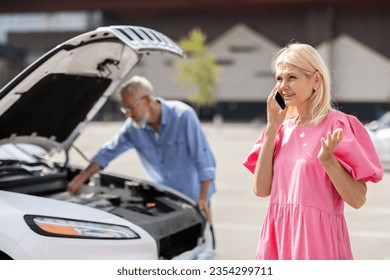 Elderly blonde woman arguing on cellphone with car insurance service on the street. Upset senior couple in road trip experience problems because of breakdown engine, old man checking auto motor - Powered by Shutterstock