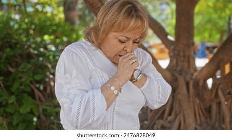 Elderly blonde woman coughing in an outdoor park setting with trees and foliage in the background, showcasing a concerned expression and wearing a white blouse and wristwatch. - Powered by Shutterstock