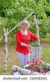 An Elderly Blonde Woman In A Red Blouse, White Breeches And A Hat Takes Care Of Flowers Planted In A Flower Bed In The Form Of A Decorative Well Made Of Birch Logs. Vertical Photo