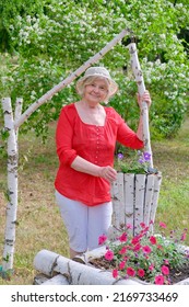 An Elderly Blonde Woman In A Red Blouse, White Breeches And A Hat Takes Care Of Flowers Planted In A Flower Bed In The Form Of A Decorative Well Made Of Birch Logs. Vertical Photo