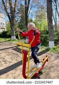 An Elderly Blonde Woman In A Red Jacket Goes In For Sports On An Exercise Bike In A Spring Park. Vertical Photo. Mirgorod, Ukraine - 05.01.2022