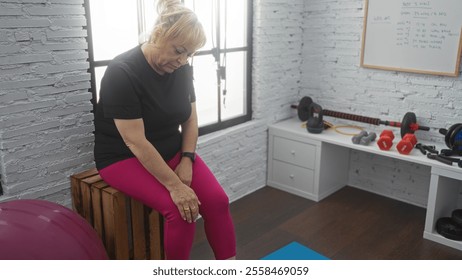 Elderly blonde woman sits resting on a wooden box in a gym with exercise equipment scattered around the room. - Powered by Shutterstock