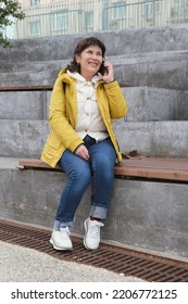 Elderly Brunette Woman Sits In A City Park On The Steps, Smiling While Talking On The Phone. Woman In A Yellow Jacket And Jeans Sits On A Bench To Talk On The Phone