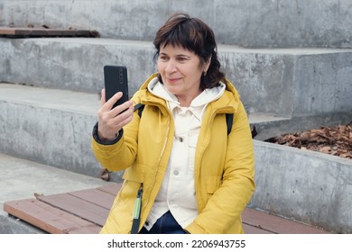 Elderly Brunette Woman Sits In A City Park On The Steps, Smiling While Talking On A Video Link. Woman Holds A Smartphone In Front Of Her. Woman In A Yellow Jacket Sits On A Bench To Talk On The Phone