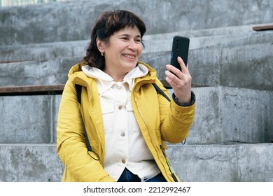 Elderly Brunette Woman Sits In A City Park On The Steps, Smiling While Talking On A Video Link. Woman Holds A Smartphone In Front Of Her. Woman In A Yellow Jacket Sits On A Bench To Talk On The Phone