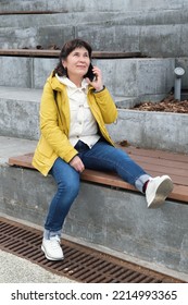 Elderly Brunette Woman Sits In A City Park On The Steps, Smiling While Talking On The Phone. Woman In A Yellow Jacket And Jeans Sits On A Bench To Talk On The Phone