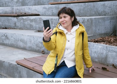 Elderly Brunette Woman Sits In A City Park On The Steps, Smiling While Talking On A Video Link. Woman Holds A Smartphone In Front Of Her. Woman In A Yellow Jacket Sits On A Bench To Talk On The Phone