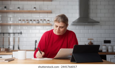 Elderly business woman sit in kitchen using tablet device, check information on document, taking notes, analyze data, looking through papers busy in paperwork - Powered by Shutterstock