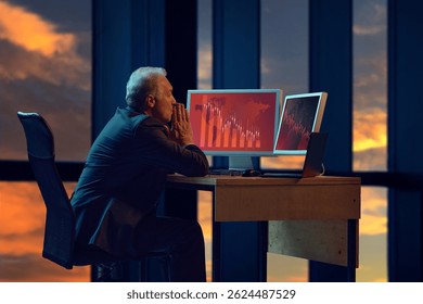 Elderly businessman in sitting late in office, looking at two computer screens displaying red graphs indicating significant downward trend. Concept of business, crisis, failure, leadership, downturn