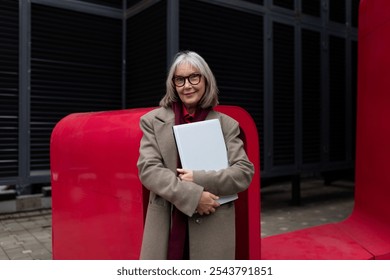 Elderly businesswoman confidently standing outside her office, holding a folder, surrounded by modern architecture - Powered by Shutterstock