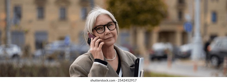 Elderly businesswoman on a phone call outside her office during a sunny afternoon in an urban setting with parked cars in the background - Powered by Shutterstock