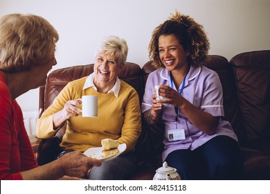 Elderly Carer Sitting With Two Of Her Patients In The Care Home. They Are Enjoying Some Cake And Tea.