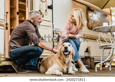 Elderly caucasian beautiful couple petting their dog together near their motorhome. Mature spouses husband and wife traveling by trailer with golden retriever
