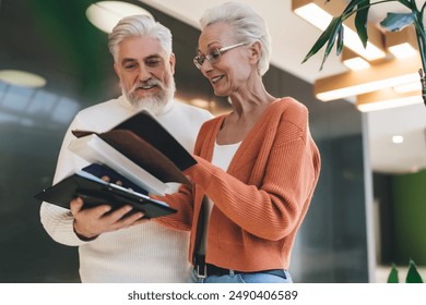 Elderly Caucasian business couple standing in modern office. Mature colleagues reviewing financial documents together, sharing work process moment, suggesting collaboration or discussing a project - Powered by Shutterstock