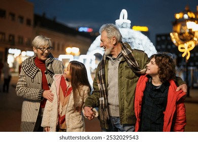 Elderly Caucasian couple delights in quality time with young boy and girl outdoors, surrounded by holiday lights. Casual winter attire and nighttime setting enhance the seasonal family joy. - Powered by Shutterstock