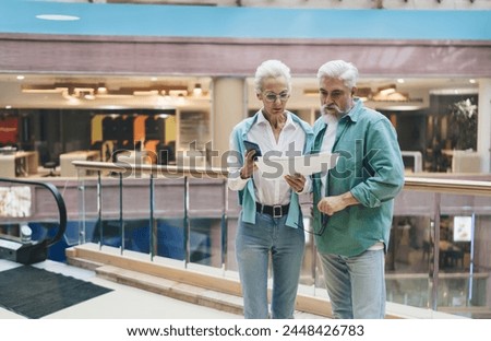 Elderly Caucasian couple discuss paperwork in a mall. Woman with phone and man with glasses show concern, implying financial or health matters. Light attire reflects a serene, spring setting.