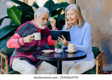 Elderly Caucasian couple sitting at outdoor cafe table sharing morning coffee while using digital tablet. Senior man and woman engaged in technology, connectivity and leisure. Warm light, green plant. - Powered by Shutterstock