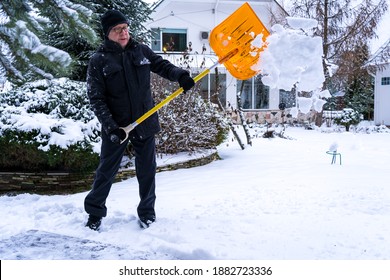 An Elderly Caucasian Man, 61 Years Old, Throws Snow With An Orange Shovel In A Garden. Healthy Lifestyle Concept In Old Age