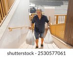 Elderly caucasian man suffers pain when carries heavy groceries bags on the exteriors stairs of the house. Stressed and ache expression. Frontal view, horizontal