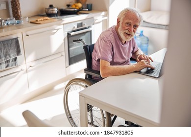 Elderly Caucasian man in the wheelchair sitting at the table in front of the laptop - Powered by Shutterstock