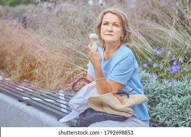 Elderly Caucasian Pretty Woman 60 Years Old Is Sitting On The Bench With Social Distancing Markup In Public Park During Coronavirus Covid-19 Epidemic And Eating Ice Cream.