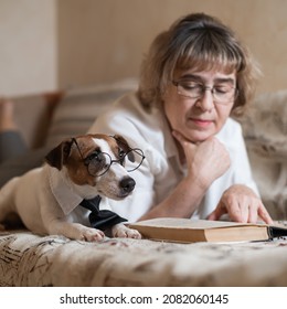 An Elderly Caucasian Woman Is Lying On A Sofa With A Smart Dog Jack Russell Terrier Wearing Glasses And A Tie And Reading A Book.