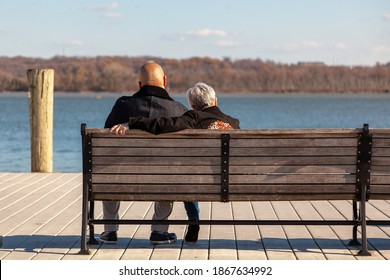 An Elderly Caucasian Woman With Short Gray Hair And A Younger African American Man With Shaved Head Are Sitting On A Bench On A Pier Looking At Sea. Image For Interracial Couple With Age Difference.