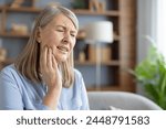 An elderly Caucasian woman sits at home, touching her cheek and grimacing in pain. She appears to be suffering from a severe toothache or jaw discomfort.