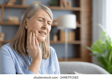 An elderly Caucasian woman sits at home, touching her cheek and grimacing in pain. She appears to be suffering from a severe toothache or jaw discomfort. - Powered by Shutterstock