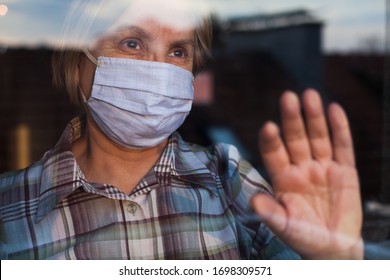 Elderly Caucasian Woman Wearing Hand Made Protective Face Mask, In Nursing Care Home, Looking Outside Window With Sadness In Her Eyes, Self Isolation Due To The Global COVID-19 Coronavirus Pandemic