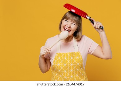 Elderly Cheerful Fun Housekeeper Housewife Woman 50s In Orange Apron Hold Spoon Cook Eat Put Frying Pan On Head Isolated Plain On Yellow Background Studio Portrait. People Household Lifestyle Concept