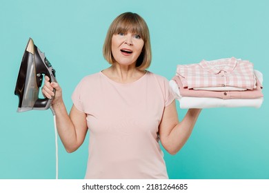 Elderly Confused Sad Housewife Woman 50s Wear Pink T-shirt Doing Housework Hold Pile Of Clothes Iron Isolated On Plain Pastel Light Blue Background Studio. Housekeeping Cleaning Tidying Up Concept.