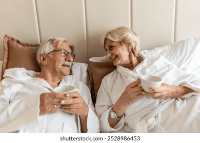 An elderly couple in bathrobes kissing each other while relaxing on a bed. They both enjoy a peaceful moment, with a cup of coffee in the morning. - Powered by Shutterstock