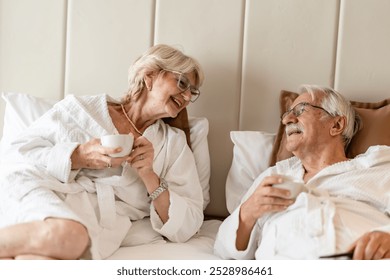 An elderly couple in bathrobes kissing each other while relaxing on a bed. They both enjoy a peaceful moment, with a cup of coffee in the morning. - Powered by Shutterstock