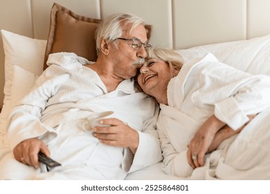 An elderly couple in bathrobes relaxes on a bed, with the man kissing the woman's forehead. They both smile, enjoying a peaceful moment, with a cup of coffee and a TV remote nearby. - Powered by Shutterstock