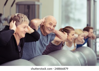 Elderly couple doing pilates class at the gym with a group of diverse younger people balancing on the gym ball with raised arms to tone their muscles in an active retirement concept