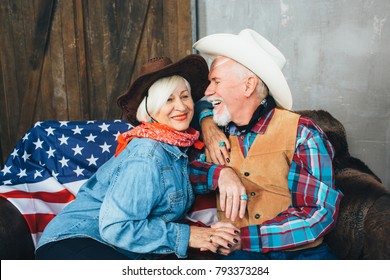 Elderly Couple, Dressed In Cowboy Hats, Laugh, Taking Each Other's Hands. Behind, On The Couch Lies The American Flag, The Celebration Of America's Independence Day