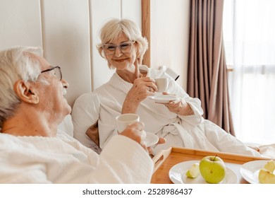 An elderly couple enjoys breakfast in bed, wearing white bathrobes, and sipping coffee. A tray with fruit and cups is placed on the bed, indicating a relaxing morning during their hotel stay. - Powered by Shutterstock