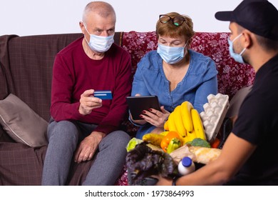 Elderly couple face mask receiving groceries from delivery man. Volunteer taking care senior family during virus outbreak. Shopping help. E payment credit card, signature tablet. High quality photo - Powered by Shutterstock