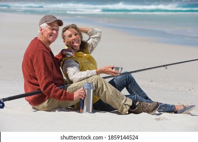 Elderly couple with a fishing rod sitting on a sunny beach and smiling at the camera while drinking coffee. - Powered by Shutterstock