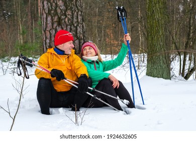 Elderly Couple Have A Rest While Walking With Nordic Walking Poles, Sitting In The Snow Under Tree In The Winter Forest. Wife And Husband Go In For Sports In Nature. Active Lifestyle Concept.