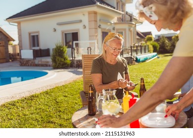 Elderly couple having friends over for an outdoor lunch in the backyard by the swimming pool, hostess setting table and having fun spending sunny summer day together
