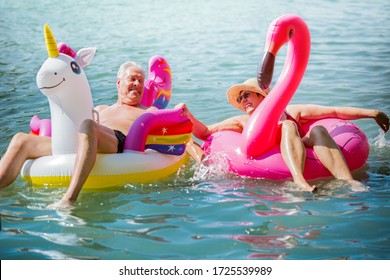 Elderly couple having fun on inflatable flamingo and unicorn. Funny active pensioners happy together enjoying summer vacation on the beach in Europe, laughing, playing the fool, splashing water. - Powered by Shutterstock