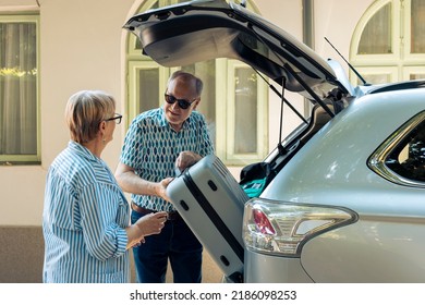 Elderly couple loading trolley in car trunk, travelling on retirement holiday vacation during summer. Senior people leaving on road trip adventure, putting suitcase and travel bags.