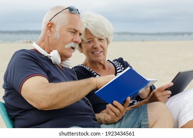 Elderly Couple Looking At Book And Tablet On The Beach