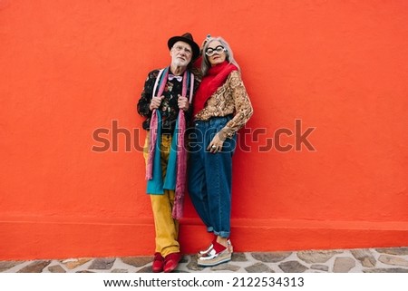 Elderly couple looking at the camera while standing together against a red background. Confident senior couple wearing colourful casual clothing outdoors. Mature couple enjoying their golden years.