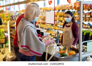 Elderly Couple In Protective Face Masks Paying At Cashdesk At Supermarket, Senior Family Shopping While Coronavirus Pandemic, Cashier Standing Behind Checkout Screen. Shopping While Quarantine Concept