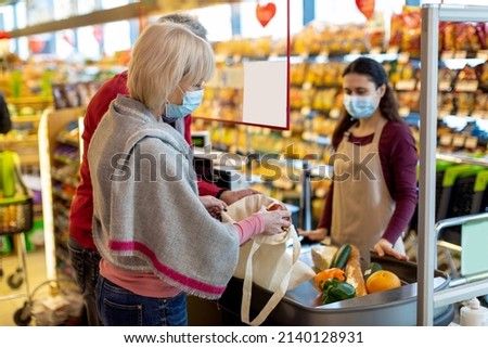 Elderly couple in protective face masks paying at cashdesk at supermarket, senior family shopping while coronavirus pandemic, cashier standing behind checkout screen. Shopping while quarantine concept