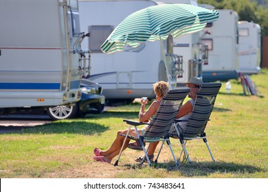 Elderly Couple Is Resting On Chairs Under A Beach Umbrella Near A Caravan